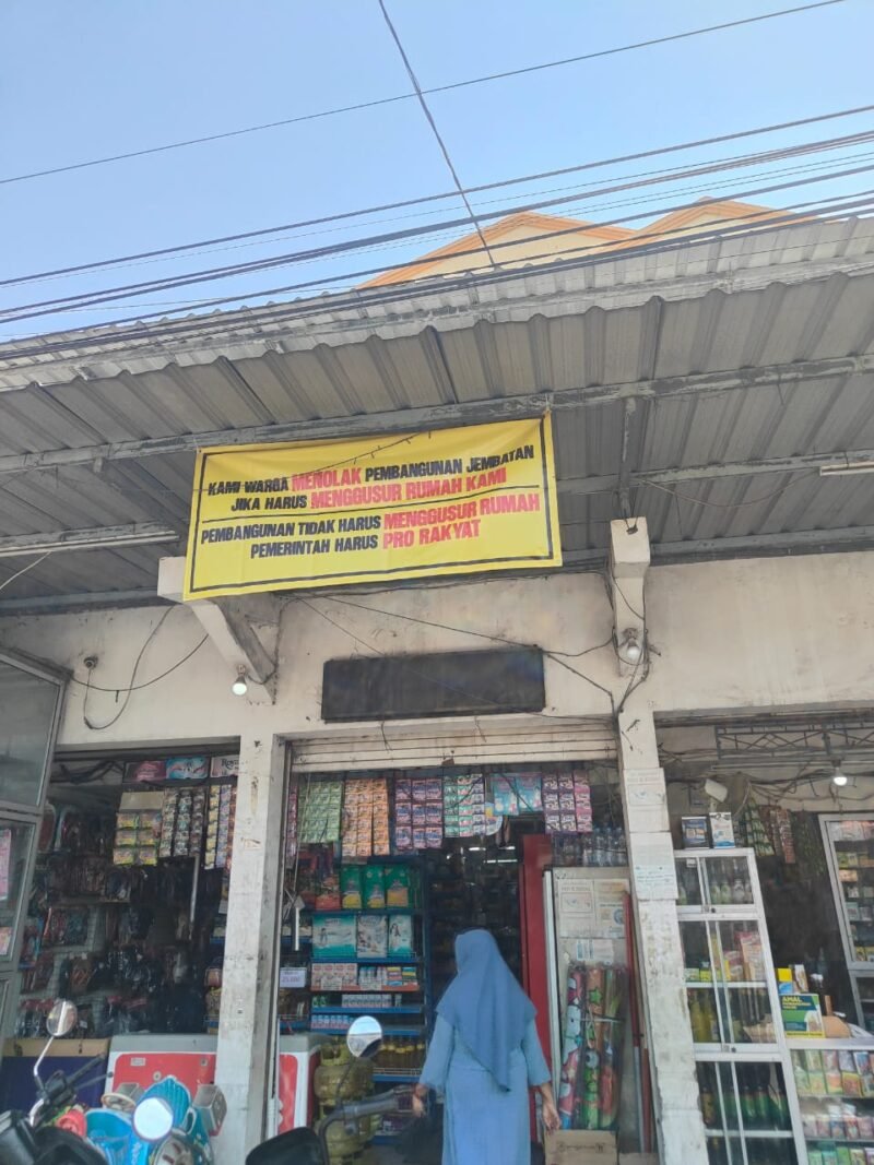 Puluhan baliho penolakan dipasang warga di sepanjang jalan utama Desa Dharma Tanjung, Sampang, sebagai bentuk protes terhadap rencana peninggian jembatan dan pelebaran jalan yang dikhawatirkan berdampak pada rumah warga.